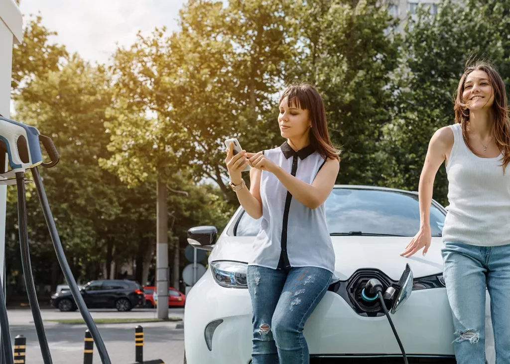 Two young woman stand in front of their car while waiting for it to charge at an EV charging station