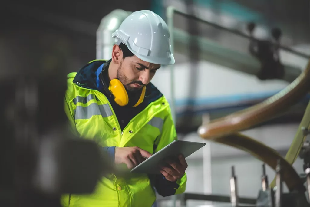 An electrician wearing a bright vest and a hard hat conducts a commercial electrical inspection with a tablet.