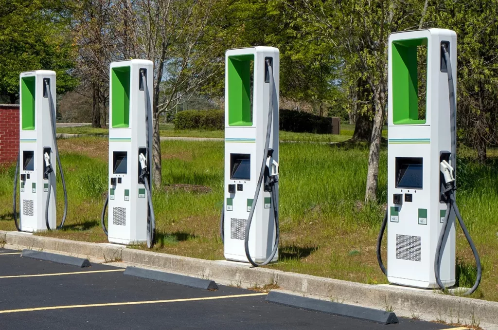 A row of white and green electric vehicle (EV) charging stations, in the parking lot of a business, with grass and trees in the background